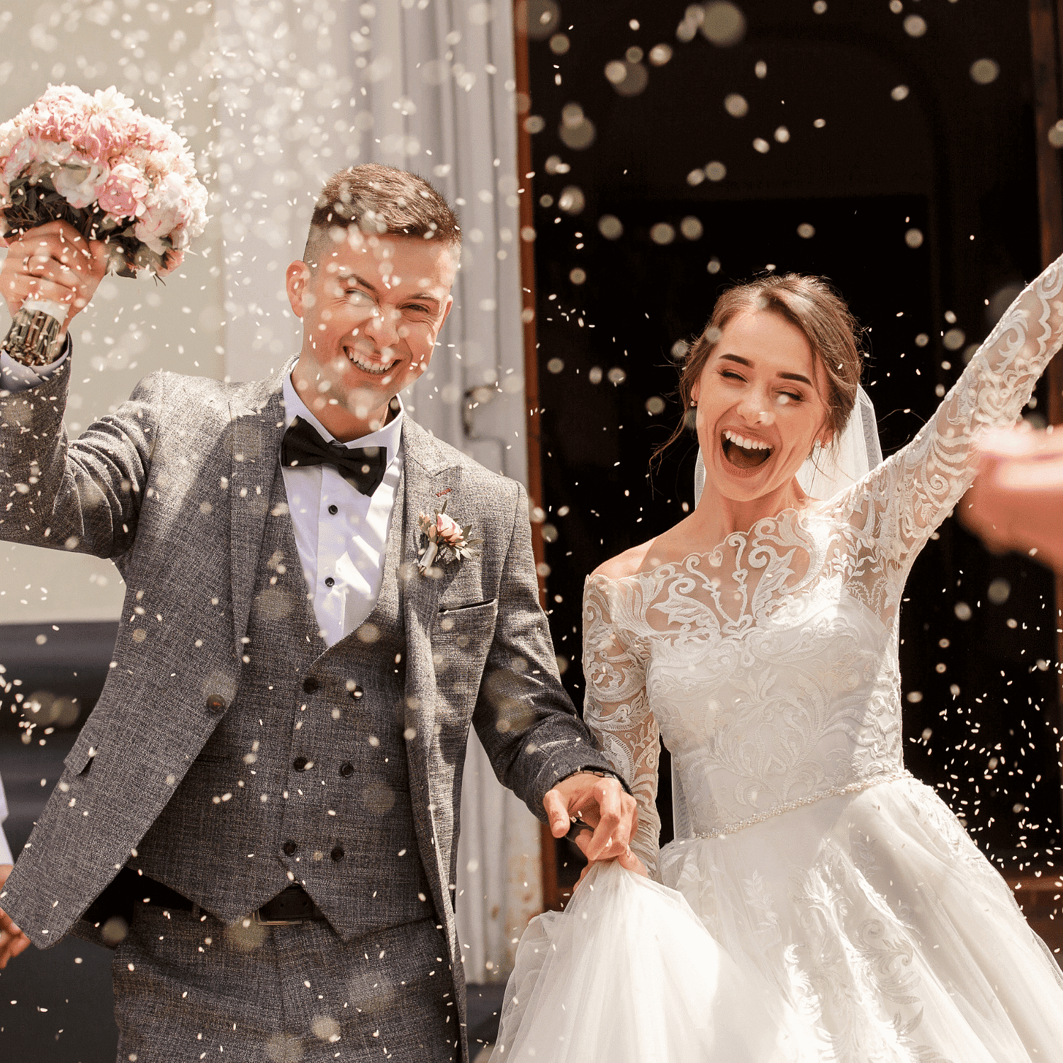 Cheerful bride and groom celebrate outside with confetti and flowers, holding hands joyfully.