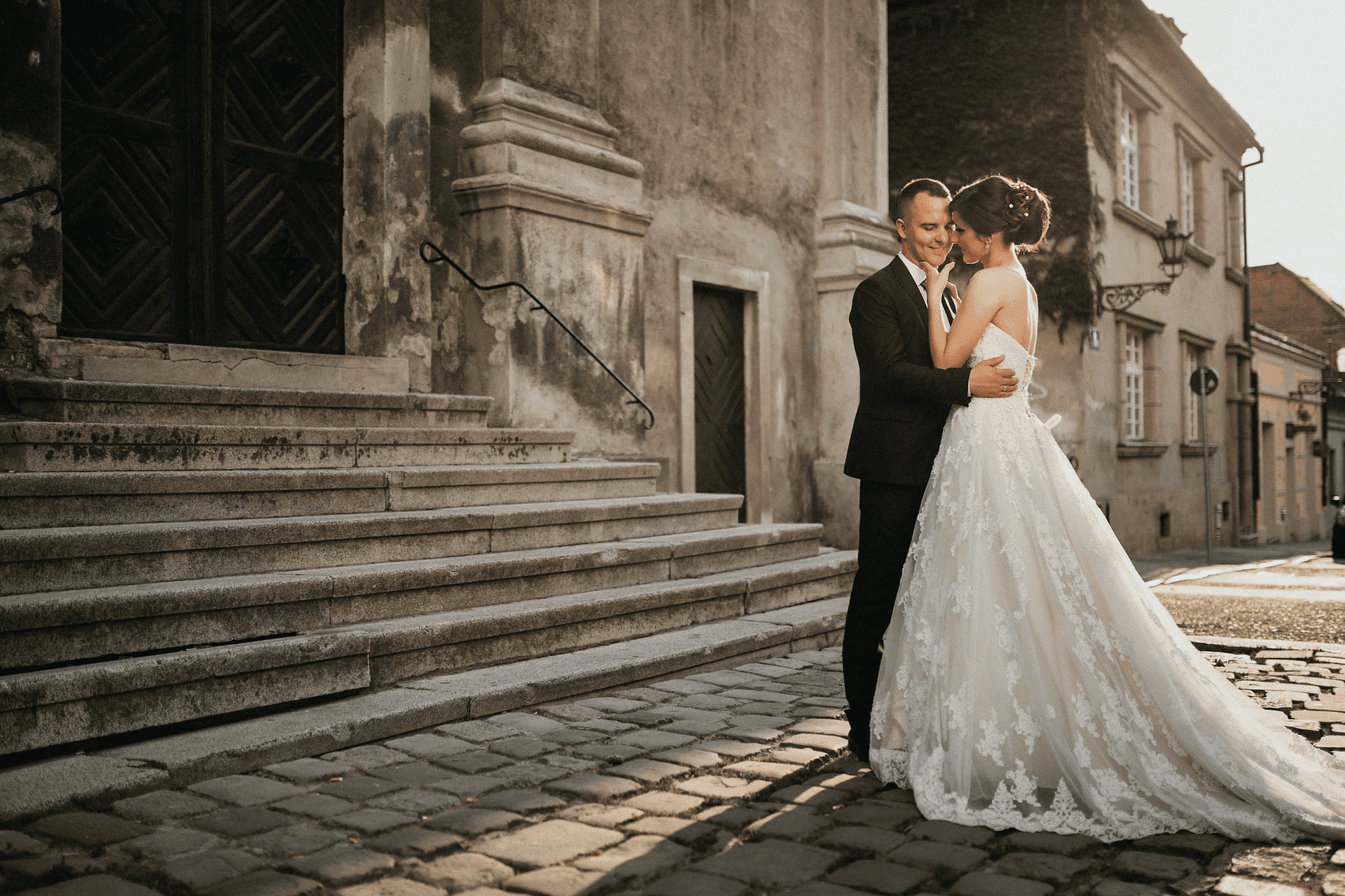 Newlywed couple shares a tender moment on a cobblestone street beside a historic building.